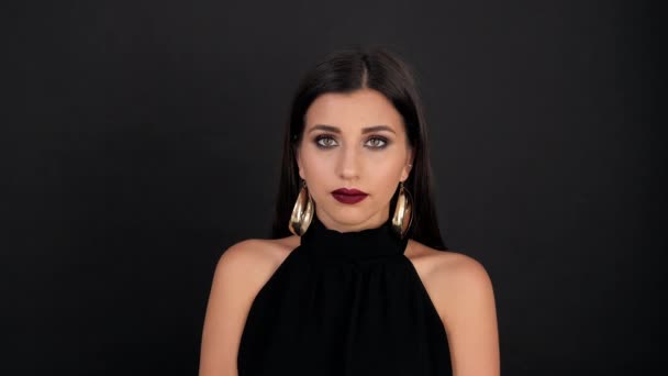 Chica con maquillaje oscuro y accesorios brillantes se sorprende de la cámara sobre un fondo negro — Vídeos de Stock