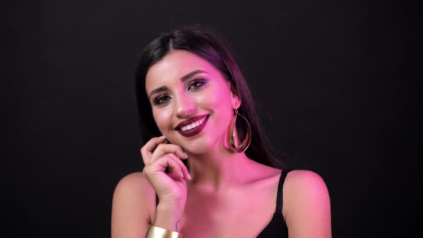 Jovencita morena con maquillaje brillante posando en la cámara con una sonrisa en la cara. Fondo negro, primer plano, luz púrpura — Vídeos de Stock