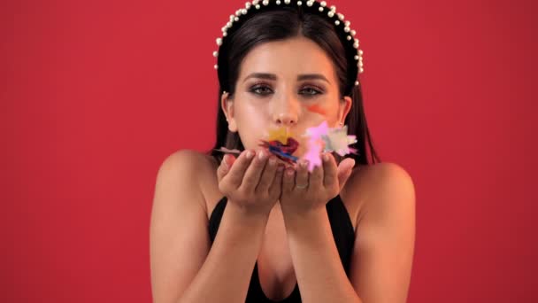 Young girl with bright holiday make-up blows confetti to the camera in the studio on a red background — Stock Video