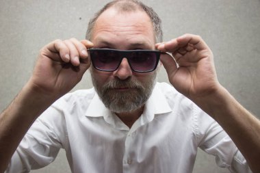 Gözlüklü sakallı gömlekli bir adam duvarın yanında duruyor..