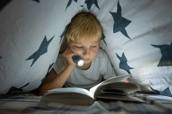 A little boy reads a book with a flashlight under the covers at night.