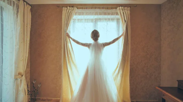 The bride opens the curtains on the window at home.