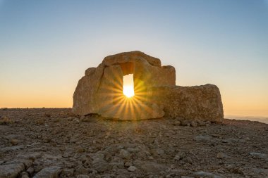 Yükselen güneş ışınları, şafak vakti, İsrail 'deki Mitzpe Ramon yakınlarındaki Judean Çölü' nün üzerindeki bir uçurumun üzerindeki halka açık bir heykel parkında, büyük bir taş bebeğin içinden geçer..