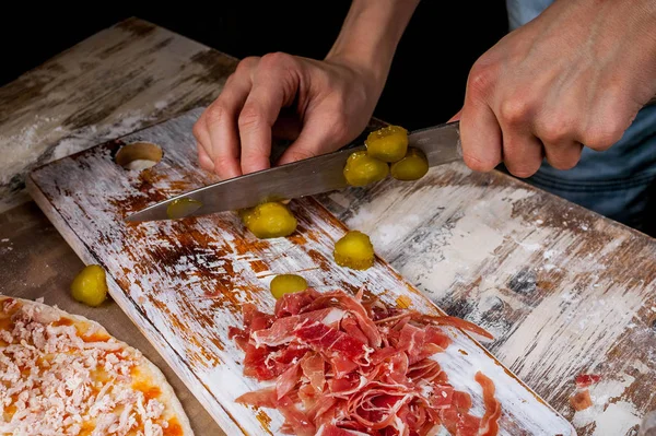 The hands of a young chef cut the vegetables and prosciutto for pizza on a wooden board. Homemade pizza with prosciutto. Close-up