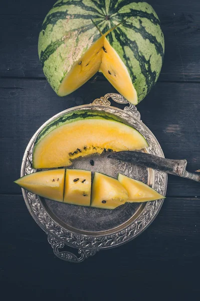 Yellow watermelon, slice of watermelon on a silver plate and knife on a dark wooden background. Vertical shot