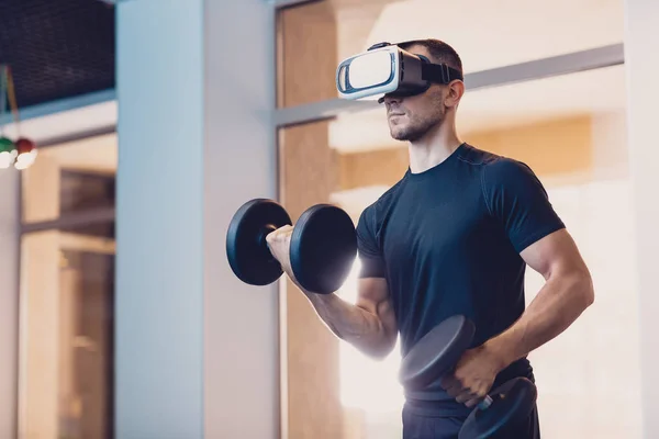 Man wearing virtual reality glasses with dumbbells. Athletic man performs an exercise on the strength of the hands with dumbbells looking into virtual reality glasses. Exercise for strength of hands