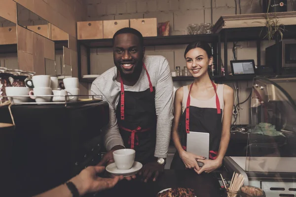 Sorridente ragazzo nero in grembiule dà tazza di caffè cotto al visitatore in caffè. Pasticceria. Barista . — Foto Stock