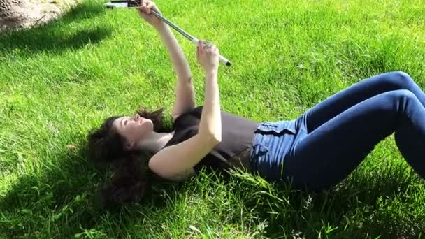 Chica acostada en la hierba en el parque haciendo selfie en el dispositivo móvil — Vídeos de Stock