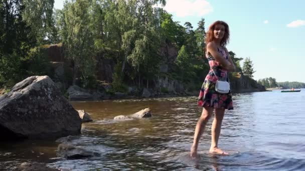 Uma menina com cabelo vermelho em um verão sundress molha os pés na água — Vídeo de Stock