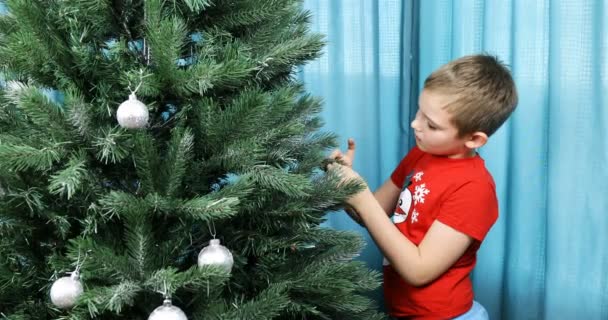 Un ragazzo in maglietta con un pupazzo di neve veste un albero di Capodanno a casa — Video Stock