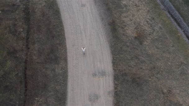 Menina em uma camisola branca monta uma bicicleta em uma estrada de terra. Vista de cima — Vídeo de Stock