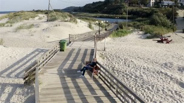 Людина контролює квадрокоптер, сидячи на лавці біля моря — стокове відео