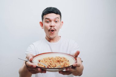 Genç bir adam bir tabak erişte ikram ederken şaşırmış görünüyor. Arkaplan düz beyaz.