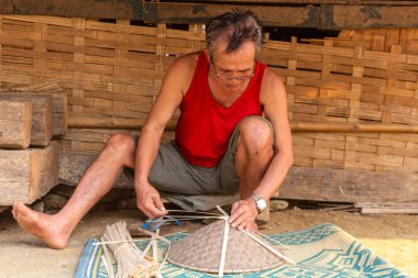 Muang Ngoi, Laos - 10 Nisan 2018: yerel adam bir el yapımı Kuzey Laos uzak bir köyde onun evinin önünde tipik Asya şapka yaptı