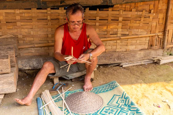 Muang Ngoi, Laos - 10 Nisan 2018: yerel adam bir el yapımı Kuzey Laos uzak bir köyde onun evinin önünde tipik Asya şapka yaptı