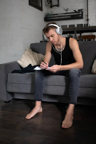 Barefoot man in headphones writing song sitting on sofa