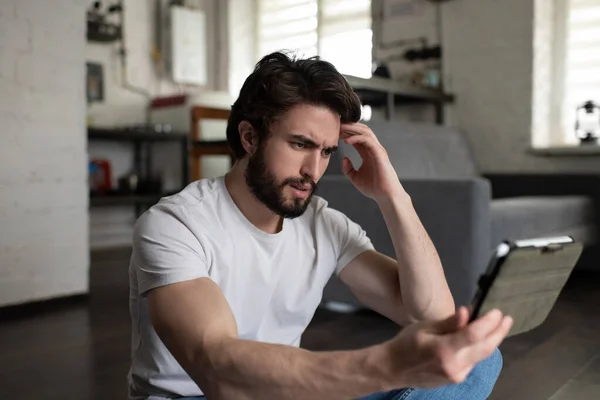 Stressed bearded man scratching head and discussing mental issues with therapist while sitting on floor and making video call on tablet