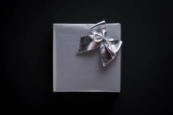 Square shape present wrapped in silver paper, decorated with silver shining bow on black background