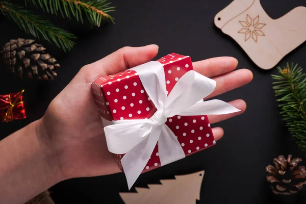 Küçük Noel hediyesi tutan el Beyaz kurdele ve yay Ladin twigs, çam kozalakları ile dekore edilmiş siyah arka plan üzerinde kırmızı puantiyeli kağıdına sarılı. Düz yatıyordu stili