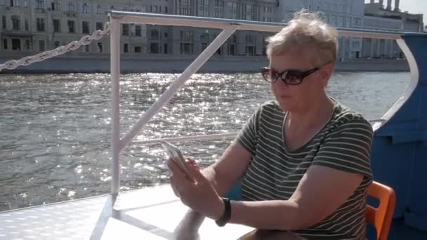 在阳光明媚的夏日里 成熟的女人坐在旅游船上 拿着后面有波光粼粼的水的智能手机 — 图库视频影像