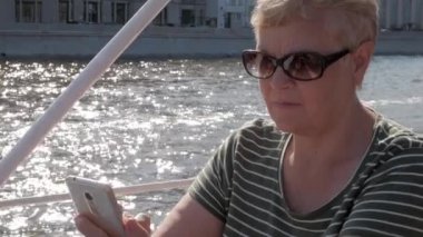 Bir teknede oturuyor ve smartphone arkasındaki köpüklü su ile güneşli yaz gününde tutan Olgun kadın kapatmak
