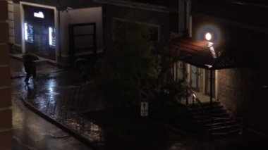 Ses ile gece yağan yağmur sırasında acele yürüyen şemsiyelerle trençkotlar kimliği belirsiz kişiler. Yüksek açılı görünüş.