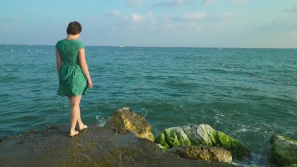 Mujer en vestido corto verde caminantes sobre rocas húmedas con salpicaduras de agua de mar a la luz del sol. Moción lenta — Vídeos de Stock