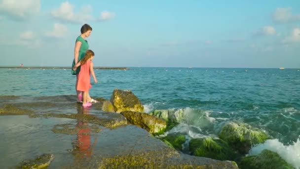 Mamá y su hija de pie sobre rocas húmedas en agua de mar con olas salpicando alrededor de divertirse. Moción lenta — Vídeos de Stock