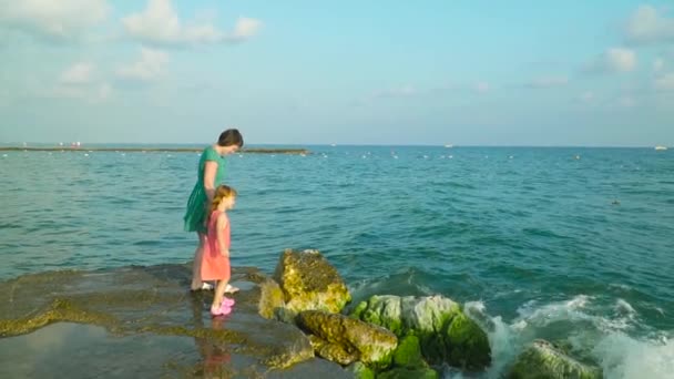 Мама и дочь стоят на мокрых камнях в морской воде с брызгами волн вокруг весело. Slow Motion — стоковое видео