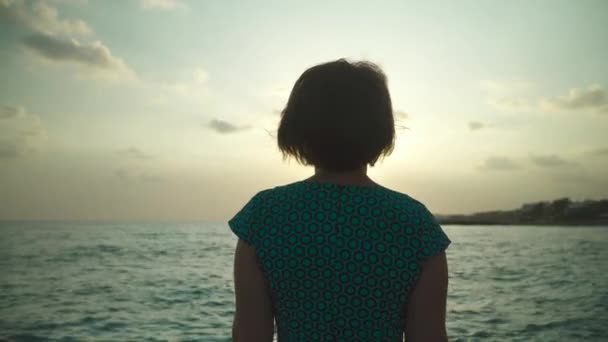 Mujer en vestido corto verde caminantes sobre rocas húmedas con salpicaduras de agua de mar a la luz del sol. En la puesta del sol — Vídeos de Stock