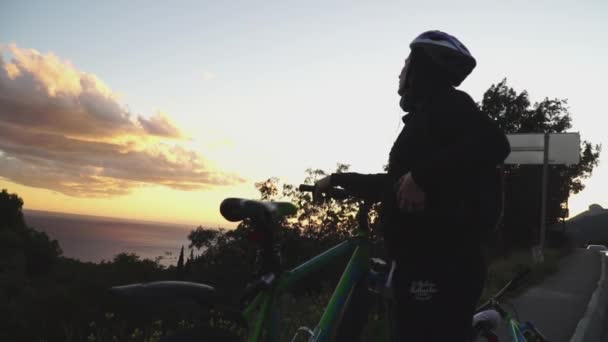 Menina com bicicleta de pé na estrada e admirando a paisagem do mar ao pôr do sol — Vídeo de Stock