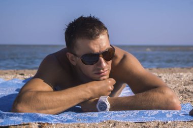 güneş gözlüğü ve beyaz saat çekici genç adam sahilde bir halı üzerinde yatıyor