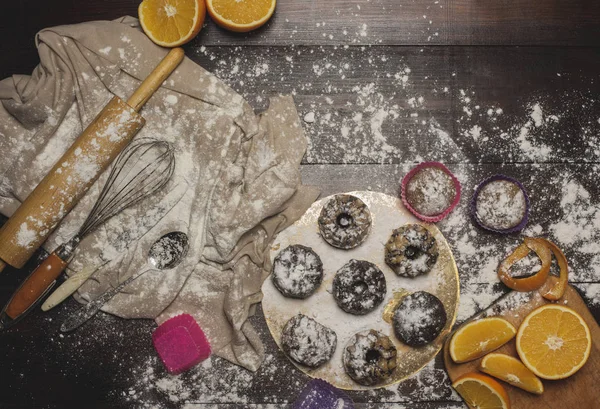 A photo of tasty cakes sprinkled with sweet powder and chocolate on a wooden brown table. Oranges and instruments  for prepare cakes. Composition with cupcakes on the table.  Top view