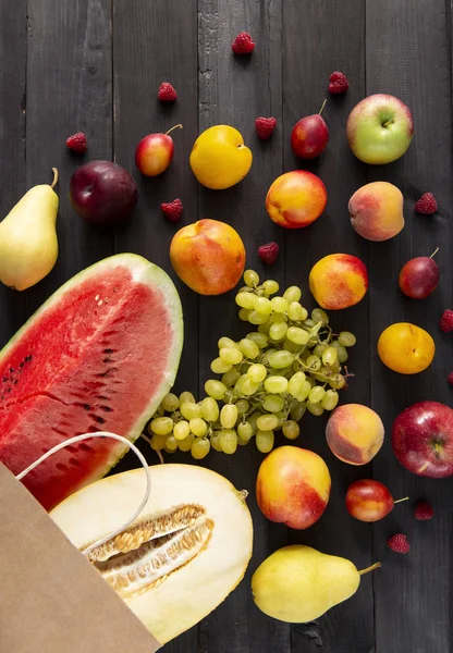 Fresh colored fruits with a paper pack on a  wooden black table. Tasty, useful, sweet set of vitamins to a dark tree. Summer and season product. Watermelon, melon, pear, plum, peach, grape. Top view