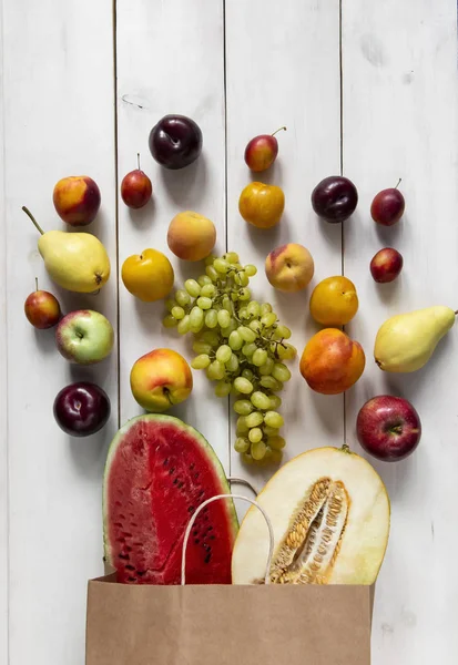 Fresh colored fruits with a paper pack on a  wooden white table. Tasty, useful, sweet set of vitamins to a light tree. Summer and season product. Watermelon, melon, pear, plum, peach, grape. Top view