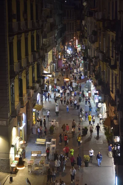 Night street life in Naples. Crowds of people walking in paved pedestrian zone between typical tall apartment buildings of Mediterranean with small shops and bars lighting street at night.