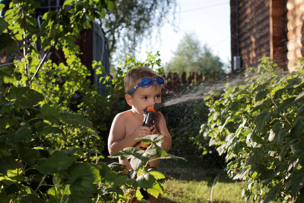 A happy little boy in the watersport goggles with a pistol in his hands watering the garden in the summer cottage during summer vacation, happy family summertime concept, outdoor lifestyle