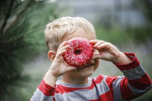Rapaz feliz comendo um donut rosa brilhante no prado — Fotografia de Stock