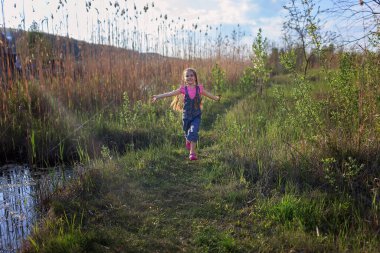 Gölün yakınındaki güzel bir çayırda koşan tatlı küçük kız, yaz tatili. Açık hava eğlencesi ve sağlıklı aktivite, insanlar tecritten sonra hayatın tadını çıkarıyor.
