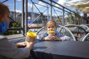 Yeni normal gerçeklik, sokak kafesinde güvende kalmak, koruyucu tıbbi maskeli çocuklar ellerinde kağıt bardaklar ve açık terasta kahvaltı için beklemek, açık hava aile yemekleri.