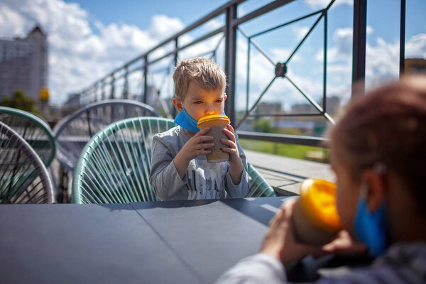 Новая нормальная реальность, безопасное пребывание в уличном кафе, дети с защитной медицинской маской держа бумажные стаканчики и ожидая завтрака на открытой террасе ресторана, открытая семейная еда
