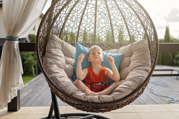 Family weekend in summer cottage, local staycation. Cute kid sitting in lotus yoga position in hanged chair on pillows near the swimming pool, happy summertime, outdoor lifestyle