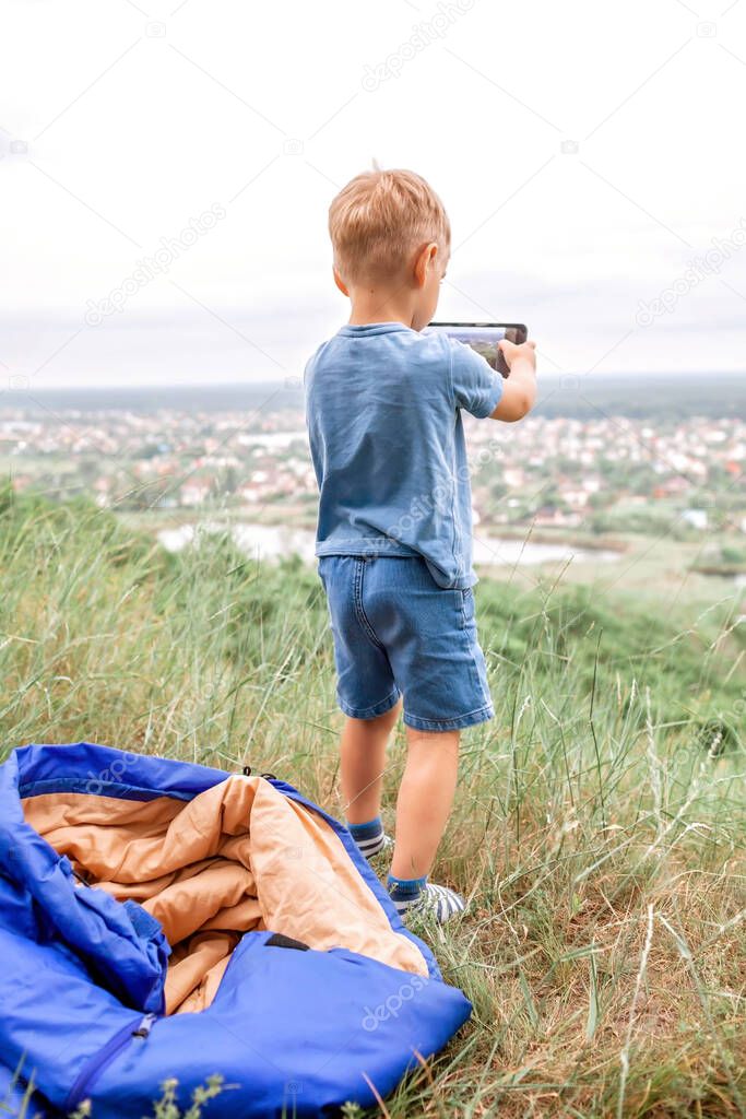 Local vacation. Cute boy having rest in sleeping bag on the top of the mountain not far from town and chatting with friends via internet, outdoor summer weekend, social distance lifestyle