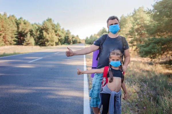 Local traveler. Middle aged father with his daughter with backpacks and wearing medical masks stopping car on the road during their local vacation, new normal summertime, active outdoor lifestyle