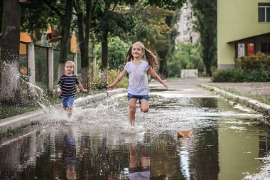 Cute happy kids jumping in the puddles after warm summer rain, outdoor walking in any weather, happy childhood and summertime, lifestyle outdoor clipart