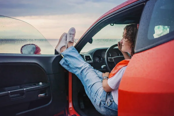 Autumn road trip, freedom and solo traveler. Pretty woman sitting in car on the bank of sea, feet on car window, looking at beautiful sunset after rain, drops on windshield, fall outdoor lifestyle