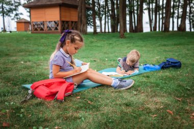 Okula geri döndüm. Salgın gerçekliğindeki açık hava dersleri sırasında yeşil çimlerin üzerinde oturan ilkokul öğrencileri. Güvenli melez eğitimi, sosyal mesafe kuralı, yeni okul rehberliği. 