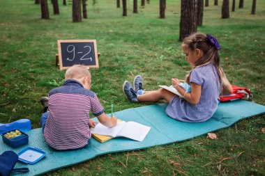Okula geri döndüm. Salgın gerçekliğindeki açık hava dersleri sırasında yeşil çimlerin üzerinde oturan ilkokul öğrencileri. Güvenli melez eğitimi, sosyal mesafe kuralı, yeni okul rehberliği. 