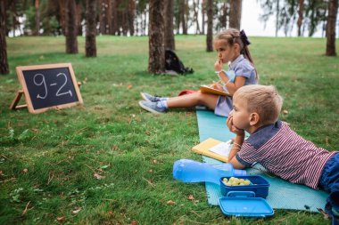Okula geri döndüm. Salgın gerçekliğindeki açık hava dersleri sırasında yeşil çimlerin üzerinde oturan ilkokul öğrencileri. Güvenli melez eğitimi, sosyal mesafe kuralı, yeni okul rehberliği. 