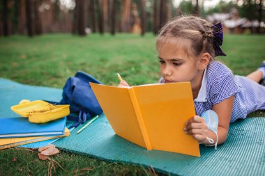 Okula geri döndüm. Salgın gerçekliğinde açık hava dersi sırasında yeşil çimlerin üzerinde oturan ilkokul öğrencisi kız. Güvenli melez eğitimi, sosyal mesafe kuralı, yeni okul rehberliği. 